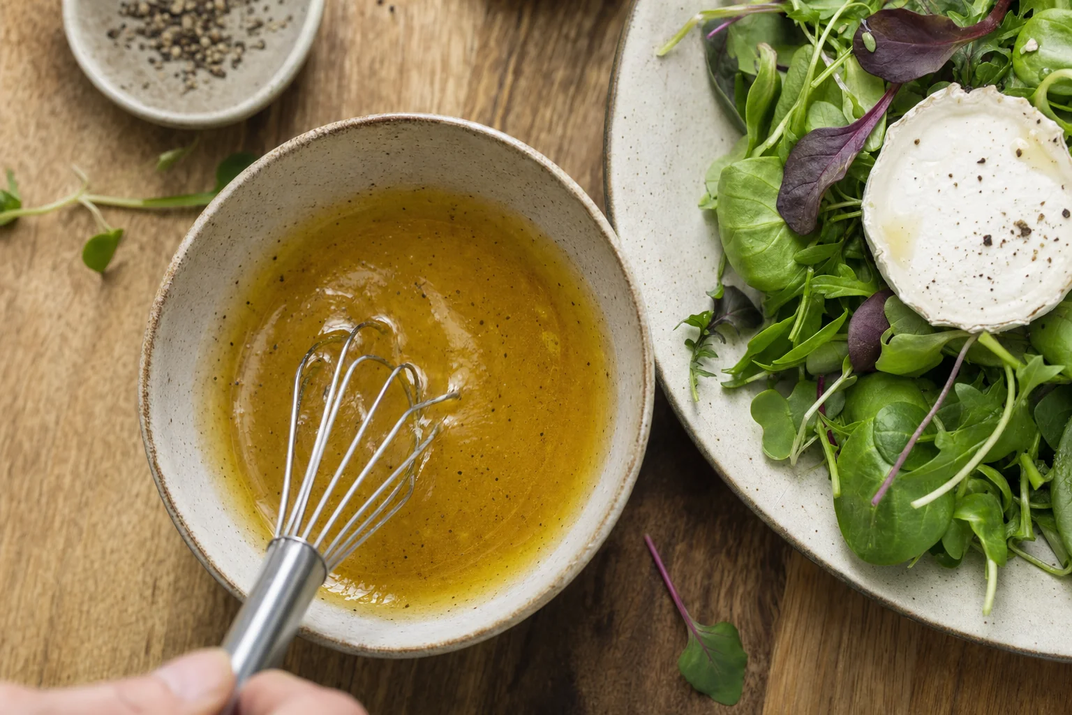 Vinaigrette dorée émulsionnée dans un bol avec un fouet, à côté d'une salade de jeunes pousses et d'un morceau de fromage de chèvre