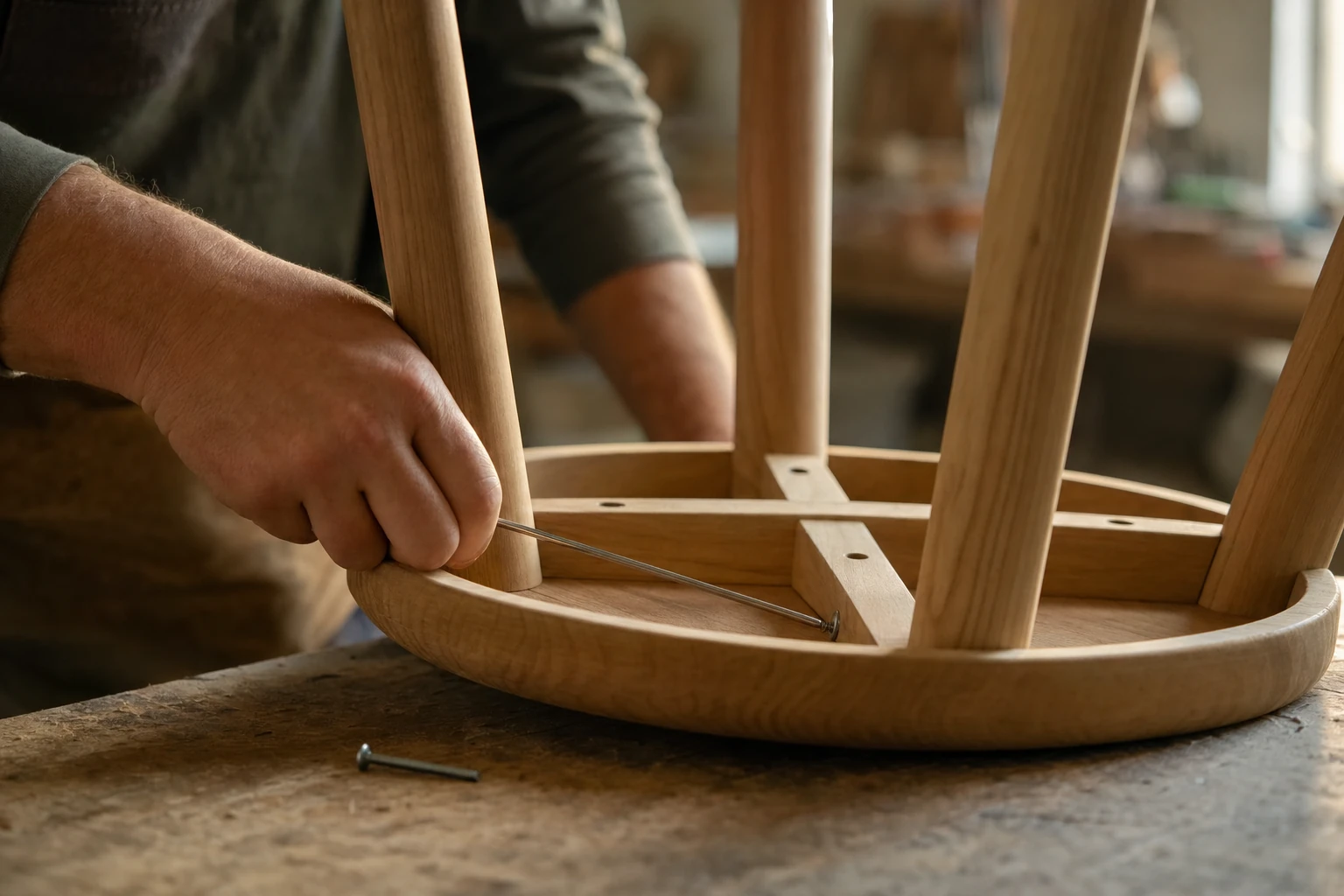 Une main d'artisan resserre une vis au centre de l'assise d'un tabouret en bois clair retourné sur un établi usé, un long tournevis fin en main, une vis de rechange posée à côté